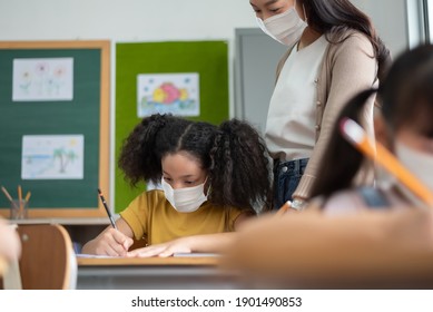 Young Asian Teacher with African-American girl wearing protective face masks in classroom at school. - Powered by Shutterstock