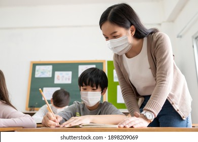 Young Asian Teacher With Little Boy Wearing Protective Face Masks Studying In Classroom During COVID-19 Pandemic. Woman Teaching Diversity Student Kids In The International School