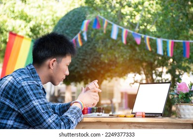 Young asian teen gay holds pink color bottle in front of laptop table, learning to draw in the park which decorated with rainbow flag and LGBTQ+ flagline, concept for special LGBT people events.