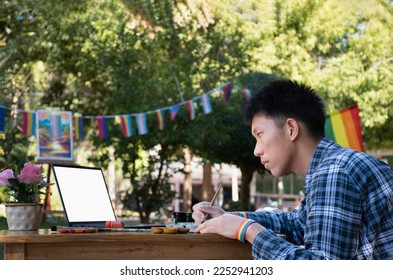 Young asian teen gay holds paintbrush and looking at laptop on table, learning to draw in the park which decorated with rainbow flag and LGBTQ+ flagline, concept for special LGBT people events.