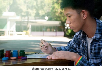 Young asian teen gay wears rainbow wristband, holds brush and drawing in the park which decorated with rainbow flag and LGBTQ+ flagline, concept for hobby of LGBT people all over the world.