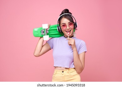 Young Asian Teenage Girl Holding Skateboard With Lollipop On Pink Background.