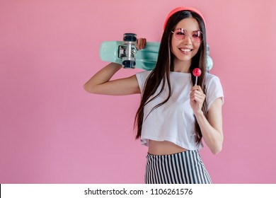 Young Asian Teenage Girl On Pink Background. Stylish Girl With Lollipop And Skateboard Isolated.