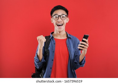 Young Asian Teenager Doing Winning Gesture Holding Mobile Phone, Happy Get Special Gift Online, Isolated On Red Background