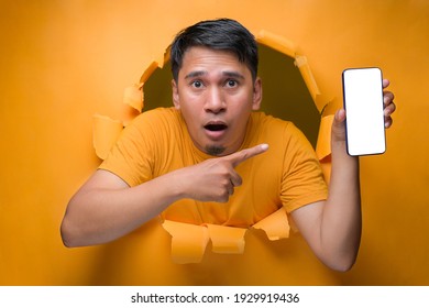 Young Asian Teenager Man With Shock Face Poses Through Torn Yellow Paper Hole, Showing Mobile Phone With Blank White Screen Display For A Copy Space. On Yellow Isolate