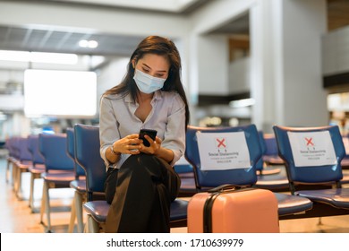 Young Asian Tourist Woman With Mask Using Phone And Sitting With Distance At The Airport