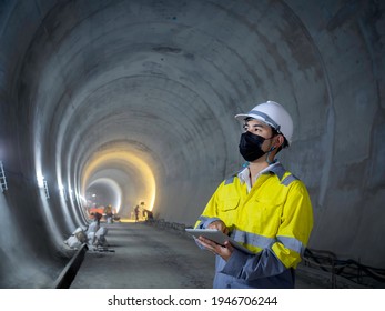 Young Asian tunnel engineering wearing high visibility jacket  face mask   white safety helmet working   using digital tablet in dark railway tunnel construction site area and copy space 