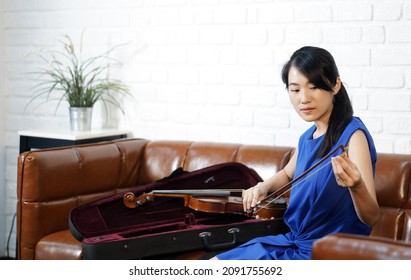 Young Asian Violinist In Blue Dress  Takes Care Of The Instrument After  Practice Session In Living Room. Rubbing Rosin On The Horse Hair Of The Violin Bow.