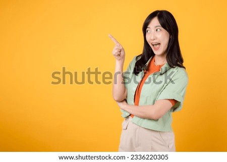 young Asian woman 30s, wearing a green shirt on an orange shirt. happy face and pointing finger to free copy space against vibrant yellow backdrop. Unlock savings and shop now!