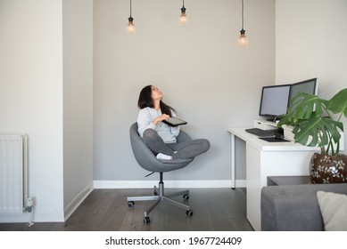 A Young Asian Woman Adjusts The Power Of The Ceiling LED Lights From Her Digital Tablet While Working From Home On Her Desktop Computer Sitting On An Office Chair In The Living Room Of A House