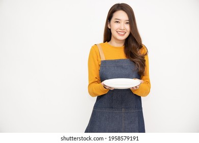 Young Asian Woman In Apron Standing And Holding Empty White Plate Or Dish Isolated On White Background