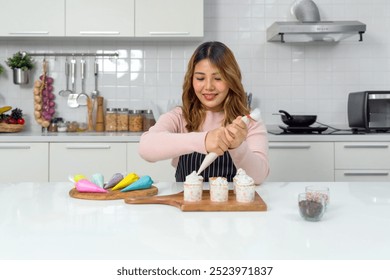 Young asian woman in an apron using a piping bag, pipe the icing onto the cupcake, decorating cupcake on a wooden board, adding a colorful touch to the cupcakes' presentation. - Powered by Shutterstock