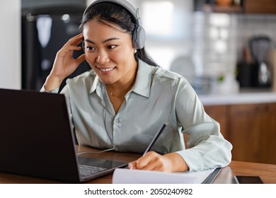 Young Asian woman attending online foreign language classes. Sitting in front of laptop computer with headphones listening course and taking notes. - Shutterstock ID 2050240982