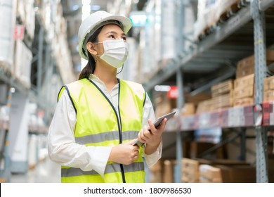Young asian woman auditor or trainee staff wears mask working during the COVID pandemic in store warehouse shipping industrial. looking up and checks the number of items store by digital tablet.