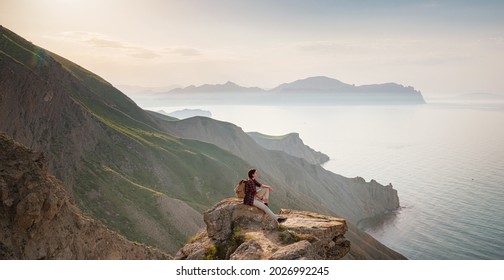 A young Asian woman with a backpack hiking in the summer sits on top of a montage and looks out over a beautiful sea bay landscape. Mountain and coastal travel, freedom and an active lifestyle