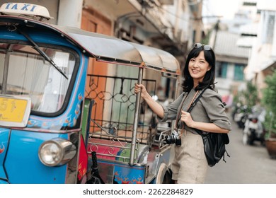 Young Asian woman backpack traveler standing a side of Tuk Tuk taxi on summer vacations at Bangkok, Thailand. Journey trip lifestyle, Asia summer tourism concept.