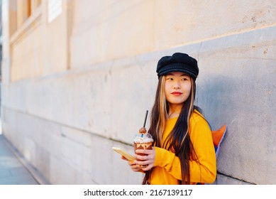 Young Asian Woman With Backpack Using Her Mobile Phone And Drinking A Chocolate Shake In The Street. Chinese Girl Doing Tourism In Europe