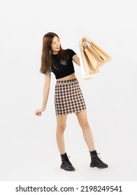 Young Asian Woman In Black Crop Top T-shirt Striped Skirt And Ankle Boots Carrying Brown Paper Shopping Bags Posing On White Background, Full Body.