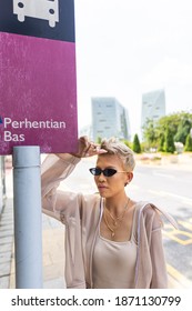 Junge Asiatische Frau mit einer schwarzen Sonnenbrille an einer Bushaltestelle auf einer Straße in Kuala Lumpur, Malaysia. Junge trendige Models mit blondem Haar und modischen Designerbekleidung auf Bushaltestelle-Schild