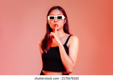 Young Asian Woman In Black Tank Top Wearing Sunglasses Posing Keep Quiet And Secret On Red Pastel Background.