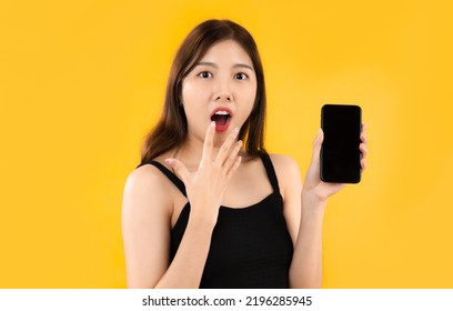 Young Asian Woman In Black Tank Top Showing Blank Black Screen Smartphone And Posing Shocked And Excited Looking Camera On Yellow Background.