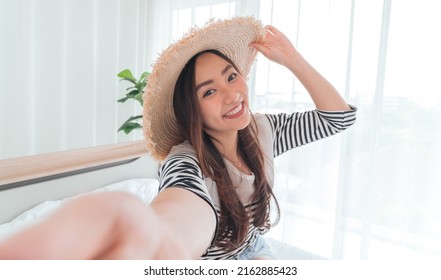 Young Asian Woman Blogger Smiling In Summer Casual Clothes And Straw Hat Talking To Facetime Video Calling With Smartphone In Bedroom. 