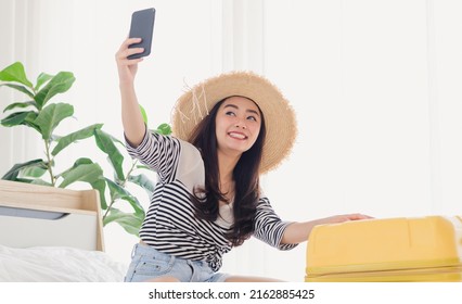 Young Asian Woman Blogger Smiling In Summer Casual Clothes And Straw Hat Talking To Facetime Video Calling With Smartphone In Bedroom