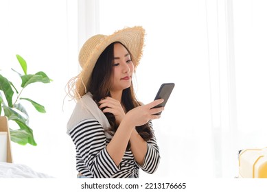 Young Asian Woman Blogger Smiling In Summer Casual Clothes And Straw Hat Talking To Facetime Video Calling With Smartphone In Bedroom