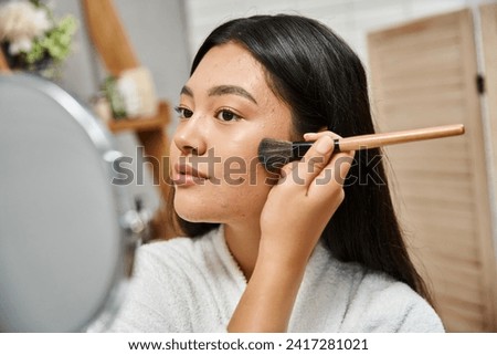 young asian woman with brunette hair and acne applying face powder and looking at mirror, skin issue