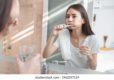 Young Asian woman brushing teeth and looking in the mirror, holding water glass, towel on the shoulder on bathroom, Concept oral hygiene and health care. - Powered by Shutterstock