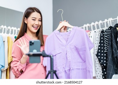 Young Asian Woman Business Owner At Fashion Store Wave Hello Smartphone Live Streaming For Sale Fashion Clothing To Customer And Present Detail On Social Media, Online Shopping Concept.