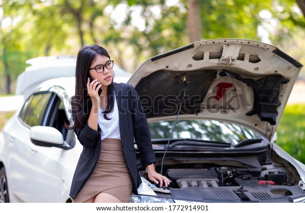 A young Asian woman is\
calling her service technician to fix a broken car on the side of\
the road, Due to an accident The engine makes it impossible to\
continue driving