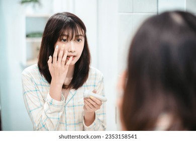 Young asian woman caring of her beautiful skin face standing near mirror in the bathroom, applying moisturizing cream on face. Smiling natural girl holding cosmetic cream or lotion - Powered by Shutterstock