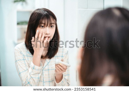 Young asian woman caring of her beautiful skin face standing near mirror in the bathroom, applying moisturizing cream on face. Smiling natural girl holding cosmetic cream or lotion