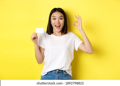 Young Asian Woman In Casual White T-shirt Showing Plastic Credit Card And Okay Gesture, Recommend Bank, Smiling At Camera, Yellow Background