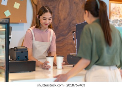 Young Asian woman coffee shop employee barista working at cafe. Smiling female waitress or cashier preparing takeaway order to customer. Small business owner and part time job working concept