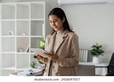 young asian woman, company worker in suit, smiling and holding digital tablet, standing over working space background - Powered by Shutterstock