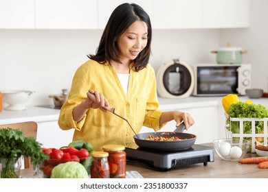 Young Asian woman cooking different vegetables in frying pan on portable electric stove at kitchen - Powered by Shutterstock