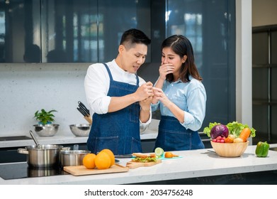 Young Asian Woman Crying In Pain Due To A Knife Cut In Finger While Chopping Vegetables In Kitchen With Husband Looking At Wound Shockingly At Home