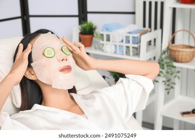 Young Asian woman with cucumber slices and facial sheet mask relaxing in spa salon, closeup