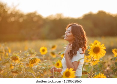 Young Asian Woman With Curly Hair In A Field Of Sunflowers At Sunset. Portrait Of A Young Beautiful Asian Woman In The Sun. Summer.