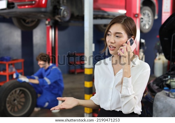 Young asian woman\
customer using smart mobile phone and calling insurance agent to\
get help or support at repair garage station with mechanic in\
uniform working blurred\
background