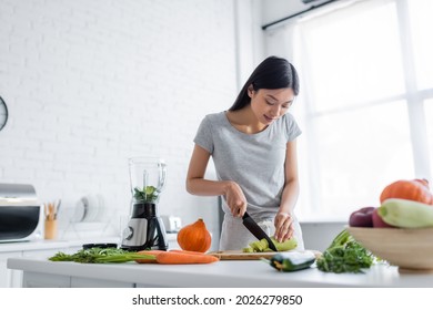 young asian woman cutting zucchini near raw vegetables and electric shaker in kitchen - Powered by Shutterstock