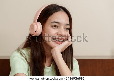 Young Asian woman with dark hair, wearing pale green top, enjoys music on pink headphones, hint of joy in smile. Music enthusiast in light-hued sweater lost in melody, pink over-ear audio gear 