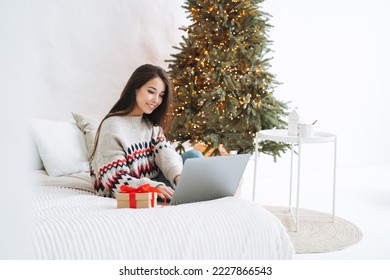 Young asian woman with dark long hair in cozy sweater using laptop on bed in room with Christmas tree at home - Powered by Shutterstock