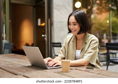 Young asian woman, digital nomad working remotely from a cafe, drinking coffee and using laptop, smiling. - Shutterstock ID 2228784891