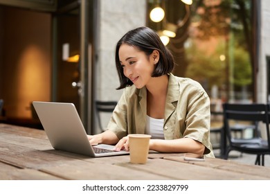 Young asian woman, digital nomad working remotely from a cafe, drinking coffee and using laptop, smiling.