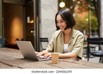 Young asian woman, digital nomad working remotely from a cafe, drinking coffee and using laptop, smiling. - Powered by Shutterstock