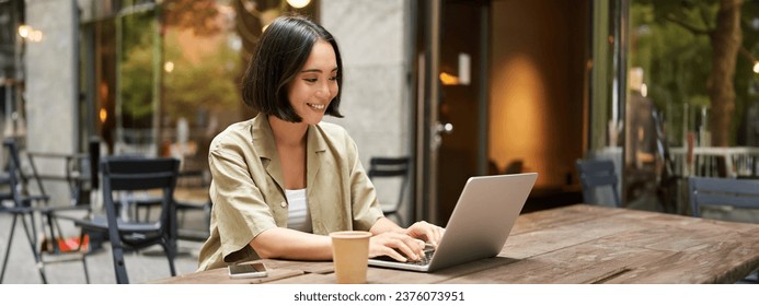 Young asian woman, digital nomad working remotely from a cafe, drinking coffee and using laptop, smiling. - Powered by Shutterstock