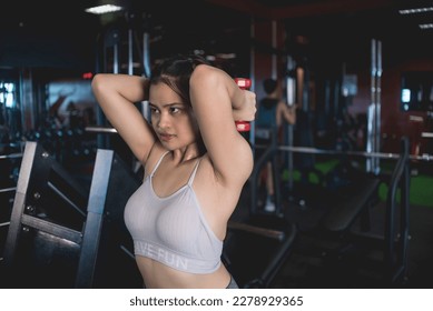 A young asian woman does standing tricep extensions with a pair of pink rubberized dumbbells. Working out triceps and arms at the gym.